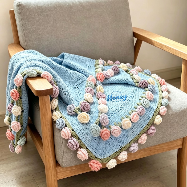 May include: A light blue crocheted blanket with a floral border of pink, white, and purple roses. The blanket is draped over a gray armchair. The word "Honey" is embroidered on the blanket.