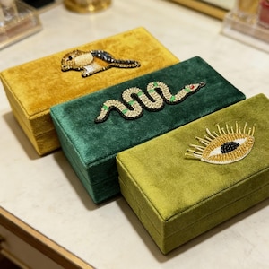 May include: Three velvet boxes in mustard yellow, emerald green, and olive green. Each box is rectangular and adorned with beaded embellishments: a bird, a snake, and an eye. The boxes are arranged on a light-colored surface.