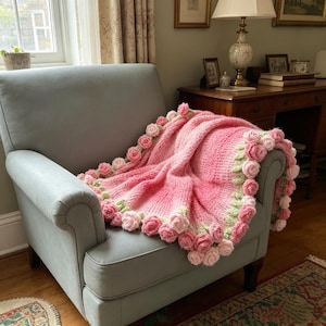 May include: A pink and rose-colored crocheted blanket with a rose border draped over a gray armchair. The blanket features a gradient of pink hues and is adorned with three-dimensional rose embellishments in shades of pink and green.