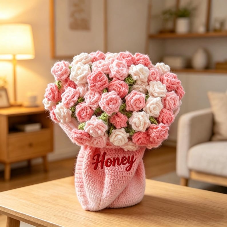May include: A handmade crochet bouquet of pink and white roses. The roses are arranged in a pink crocheted vase with the word "Honey" in red script. The bouquet is displayed on a wooden table.