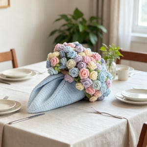 May include: A light blue crocheted napkin holder shaped like a cone, adorned with a bouquet of pastel-colored crocheted roses. The roses are in shades of pink, blue, yellow, and purple. The napkin holder is on a white tablecloth.