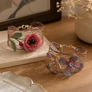 Peut inclure: Deux bracelets manchette en maille métallique brune. L'un présente une grande fleur en tissu rouge avec des feuilles vertes. Le second bracelet arbore deux fleurs plus petites aux tons dorés et violets, chacune ornée d'un petit cristal bleu.