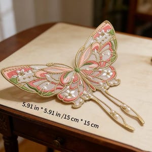 Puede incluir: Accesorio para el cabello con forma de mariposa bordada, de 15 cm por 15 cm. La mariposa presenta bordados rosas, verdes y dorados con detalles de perlas. El accesorio está diseñado con dos largas púas doradas.