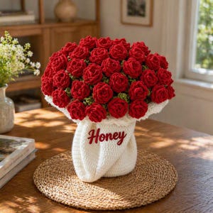 May include: A handcrafted bouquet of red crochet roses in a white crocheted vase. The word "Honey" is written in red script on the vase. The bouquet sits on a round, woven placemat on a wooden table.