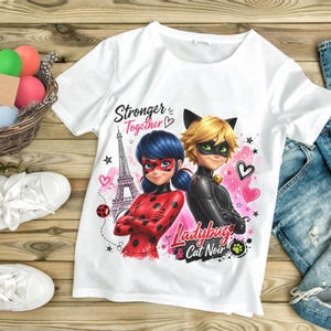 May include: White t-shirt featuring a colorful graphic of Ladybug and Cat Noir from Miraculous: Tales of Ladybug & Cat Noir. The design includes the Eiffel Tower, hearts, and the text "Stronger Together" and "Ladybug & Cat Noir".