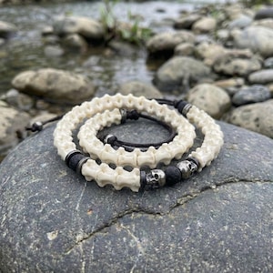 Op de afbeelding: Een gelaagde ketting- en armbandset met botvormige kralen, zwarte lavakralen en zilveren schedelaccenten. De sieraden worden getoond op een grijze steen, met een vage rivier en rotsen op de achtergrond.