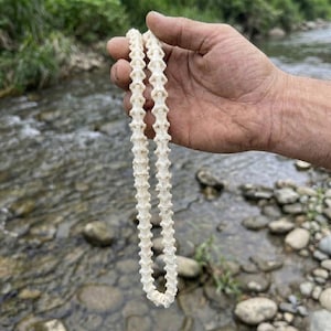Op de afbeelding: Een cr&egrave;mekleurige ketting gemaakt van kleine, botachtige segmenten. De ketting wordt in een hand gehouden, met een vage achtergrond van een rivier en groen.