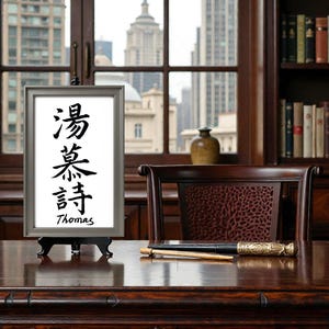 May include: A framed artwork with Chinese calligraphy and the name "Thomas" displayed on a wooden desk. The desk is in front of a window overlooking a cityscape. A decorative writing instrument rests on the desk.
