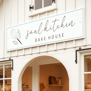 May include: A white storefront with a rectangular sign reading soul kitchen BAKE HOUSE in black text with a whisk icon. The shop features large glass windows displaying various loaves of bread and baked goods on shelves inside the bakery.