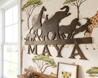 May include: A dark brown wooden wall sign featuring silhouettes of a giraffe, elephant, zebra, and antelope above the name MAYA in capital letters. The sign is mounted on a wall with a safari-themed pattern featuring trees and animals.