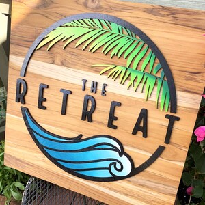 May include: A wooden sign with a black circle containing a green palm tree and a blue wave. The text "THE RETREAT" is in black letters.