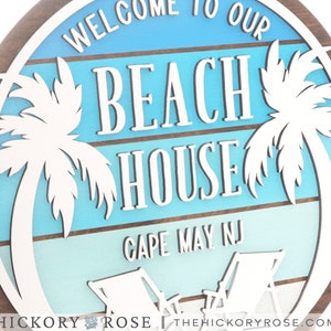 May include: Welcome to our Beach House sign with a blue and white ombre background, palm trees, and beach chairs. The sign also includes the text "Cape May NJ".