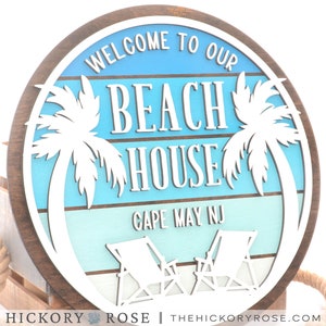 May include: A round wooden sign with a blue and white beach scene. The sign says "Welcome to our Beach House Cape May NJ" with palm trees and beach chairs.