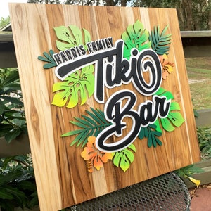 May include: A wooden sign with the text "Harris Family Tiki Bar" in black and white lettering. The sign is decorated with green and yellow tropical leaves and flowers.