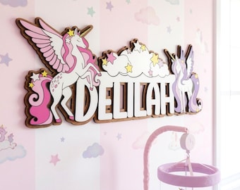 Op de afbeelding: Houten wanddecoratie met de naam "DELILAH" en eenhoorn- en wolkenontwerpen. Het kunstwerk is in roze, wit en bruin, met steraccenten. De achtergrond is een roze gestreepte muur met eenhoorn- en sterpatronen.
