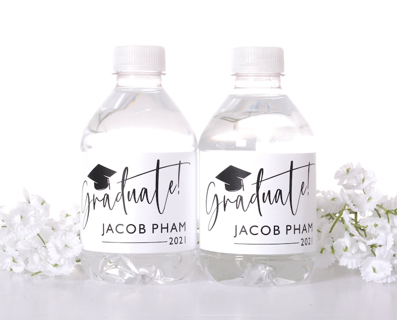 May include: Two clear plastic water bottles with white labels that say "Graduate!" in black script with a graduation cap icon. The labels also include the name "Jacob Pham" and the year "2021".