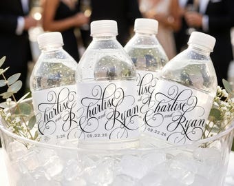 Op de afbeelding: Doorzichtige plastic waterflessen met witte etiketten voorzien van zwarte decoratieve sierletters met de tekst "Charlise and Ryan" gevolgd door de datum "09.22.26". De flessen zijn gevuld met water en staan in een kom gevuld met ijsblokjes.