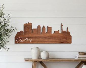 Op de afbeelding: Houten wandkunst met de skyline van Lynchburg, Virginia. De stadsgezicht is uit één stuk hout gesneden en toont verschillende gebouw silhouetten. Het woord "Lynchburg" is in cursief geschreven. De kunst is bevestigd aan een witte wand met lamellen.