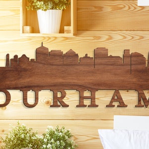 May include: A wooden wall art piece featuring the Durham, North Carolina skyline silhouette. The dark brown artwork is mounted on a light wood background. The city name, "DURHAM," is cut out below the skyline.