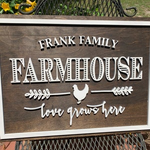 May include: A wooden farmhouse sign with white lettering that reads "Frank Family Farmhouse - Love Grows Here". The sign has a white border and a white silhouette of a chicken with a wheat stalk on either side.