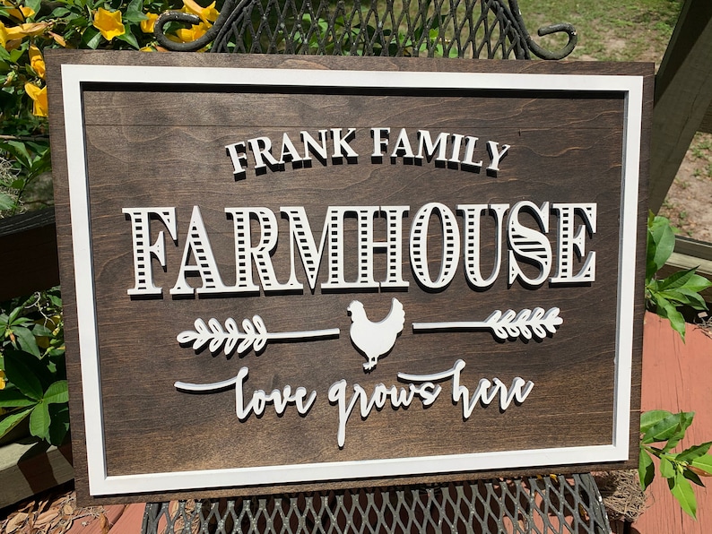 May include: A wooden farmhouse sign with white lettering that reads "Frank Family Farmhouse - Love Grows Here". The sign has a white border and a white silhouette of a chicken with a wheat stalk on either side.