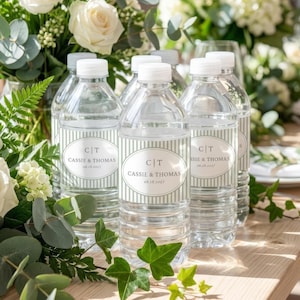 May include: Several clear plastic water bottles with white caps are arranged on a wooden surface. Each bottle has a label with the text "CT CASSIE & THOMAS 06.18.2027" in a white oval shape, surrounded by light green stripes. The background includes white flowers and greenery.