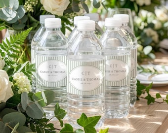 Op de afbeelding: Een aantal doorzichtige plastic waterflessen met witte doppen. Elke fles heeft een etiket met de tekst "CT CASSIE & THOMAS 06.18.2027" in een witte ovaal, omringd door lichtgroene strepen. De achtergrond toont witte bloemen en groen.