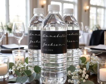 Op de afbeelding: Drie doorzichtige plastic waterflessen met witte doppen, elk voorzien van een zwart rechthoekig etiket. Het etiket toont de namen Annabelle en Jackson in een wit handschrift, met de datum 12.02.2024 in een schreefloos lettertype.