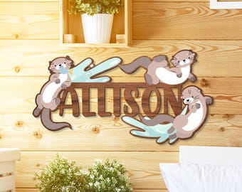 Op de afbeelding: Houten wandkunst met de naam "ALLISON" in bruine letters, omringd door otterillustraties. De otters zijn in bruin en wit, met lichtblauwe accenten. Een charmant, gepersonaliseerd decoratief item.