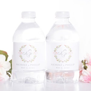 May include: Two clear plastic water bottles with white labels. The labels feature a floral wreath with the initials "N & P" in a gray script font. The text "Nichole & Phillip" and "May 5, 2021" is printed below the wreath. The bottles are surrounded by pink flowers.