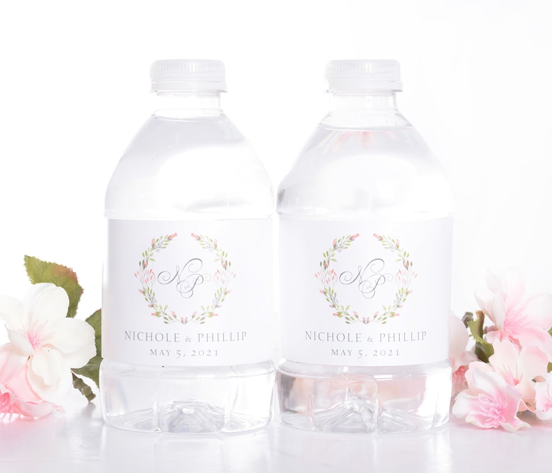 May include: Two clear plastic water bottles with white labels. The labels feature a floral wreath with the initials "N & P" in a gray script font. The text "Nichole & Phillip" and "May 5, 2021" is printed below the wreath. The bottles are surrounded by pink flowers.