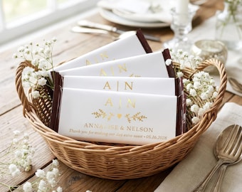 Op de afbeelding: Een gevlochten rieten mand gevuld met chocoladerepen verpakt in wit papier met goudfolie letters. De tekst luidt A | N, Annalise & Nelson, en Thank you for making our day extra sweet 05.16.2028. Kleine witte bloemetjes versieren de mand.