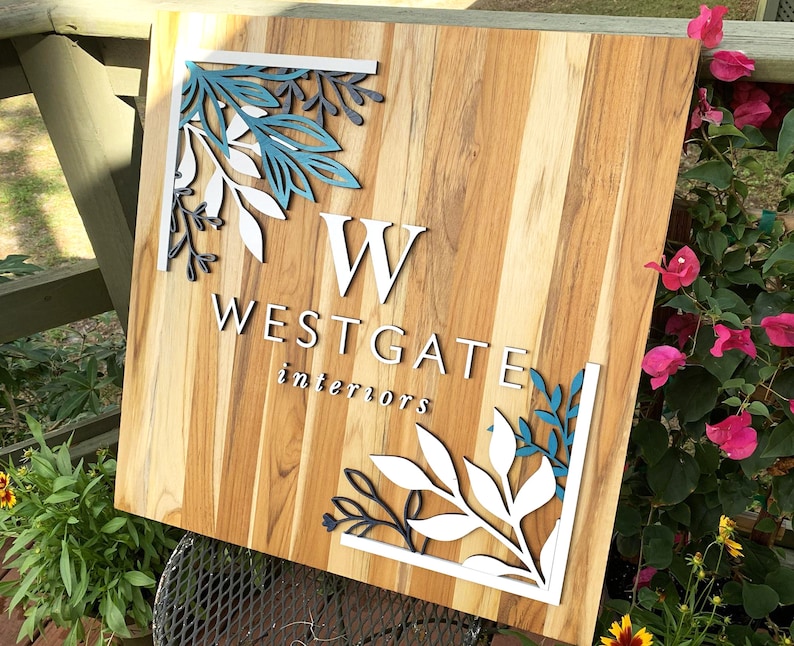 May include: A wooden sign with a white background and a teal and black floral design. The sign says "W Westgate interiors" in white letters.