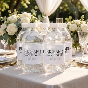 May include: Clear plastic water bottles with white labels that read "RICHARD and GRACE 04.18.2027" are arranged on a table. The bottles are surrounded by white roses and greenery, suggesting a wedding or event setting.