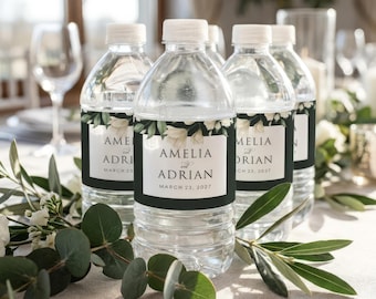 Op de afbeelding: Doorzichtige plastic waterflessen met witte doppen, elk met een etiket met de namen "Amelia en Adrian" en de datum "23 maart 2027". De etiketten hebben een donkergroene rand en bloemmotieven. De flessen staan op een tafel met groen.