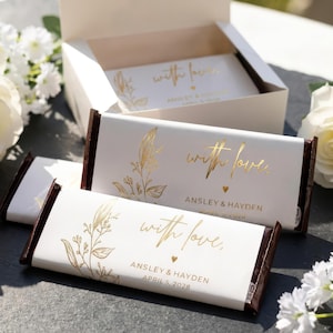 May include: White chocolate bar wrappers featuring gold foil botanical illustrations and the text "with love," "ANSLEY & HAYDEN," and "APRIL 5, 2028." The wrappers are displayed on chocolate bars and inside an open white cardboard gift box.