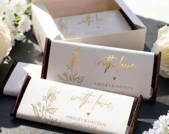 Op de afbeelding: Witte chocoladereepverpakkingen met botanische illustraties in goudfolie en de tekst "with love,", "ANSLEY & HAYDEN" en "APRIL 5, 2028". De verpakkingen liggen op chocoladerepen en in een open witte geschenkdoos.