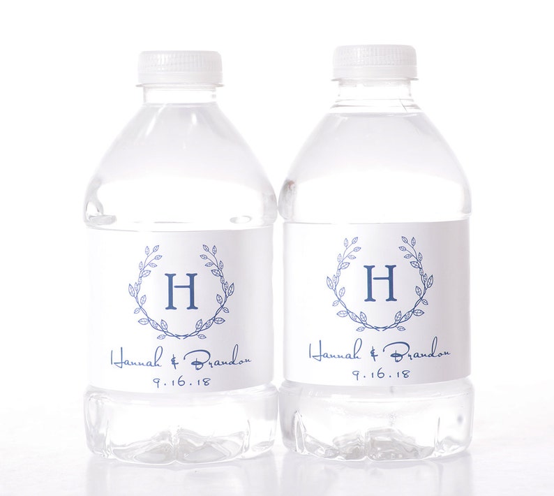 May include: Two clear plastic water bottles with white labels. The labels feature a blue wreath with the letter "H" in the center. The text "Hannah & Brandon 9.16.18" is printed below the wreath.