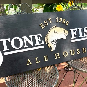 May include: A black wooden sign with white and gold lettering. The sign reads "Stone Fish Ale House Est 1980". The sign has a stylized fish and a letter "H" carved into it.
