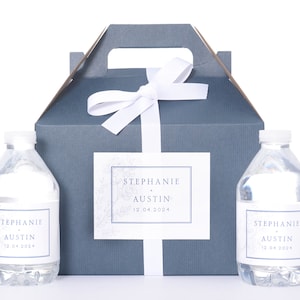 May include: Two clear plastic water bottles with personalized labels that say "Stephanie + Austin 12.04.2024". The bottles are in front of a gray gift box with a white ribbon and a matching label.