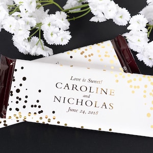 May include: White chocolate bar wrapper with gold confetti and black polka dots. The wrapper has the text "Love is Sweet! Caroline and Nicholas June 24, 2018".