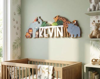 Op de afbeelding: Een houten wanddecoratie met de naam "KEVIN" met een junglethema. De letters zijn wit met een houten rand, en het ontwerp bevat kleurrijke dierenillustraties zoals een leeuw, een olifant, een giraffe en een nijlpaard. De decoratie is boven een wieg geplaatst.