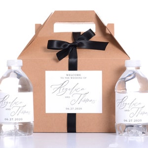 May include: Two clear plastic water bottles with white labels that say "Angelica and Thomas 06.27.2020". The bottles are in front of a brown cardboard box with a black ribbon and a white label that says "Welcome to the wedding of Angelica and Thomas 06.27.2020".