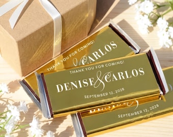 Op de afbeelding: Een bruine kartonnen geschenkdoos met een witte strik en verschillende rechthoekige chocoladerepen verpakt in glanzend goudfolie. De labels bevatten de witte tekst "THANK YOU FOR COMING!", "DENISE & CARLOS" en "SEPTEMBER 12, 2028" op de voorzijde.