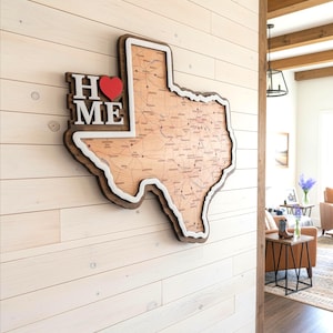 May include: A wooden wall art piece shaped like the state of Texas, featuring a detailed map design. The artwork includes the words "HOME" with a red heart, and a white border. The piece is mounted on a white wooden wall.