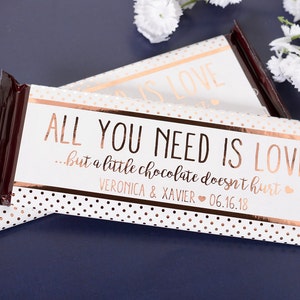 May include: A white chocolate bar wrapper with gold foil accents and polka dots. The wrapper has the text "ALL YOU NEED IS LOVE...but a little chocolate doesn't hurt." and "VERONICA & XAVIER 06.16.18".