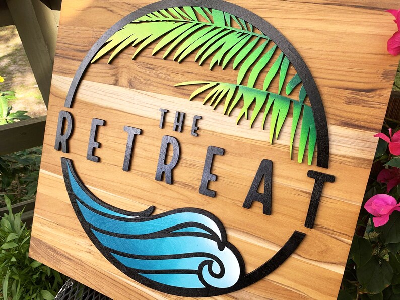 May include: A wooden sign with a black circle containing a green palm tree and the text "THE RETREAT" in black letters. A blue wave with a white swirl is below the text.