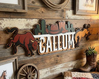 Op de afbeelding: Een houten bord met de naam "CALUM" in witte letters, met een cowboythema. Het bord bevat een stier, een paard, een cowboyhoed en cactussen. De achtergrond is een rustieke houten muur.