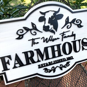 May include: White farmhouse sign with black lettering and a black silhouette of a cow. The sign reads "The Wilson Family Farmhouse Established 2012".