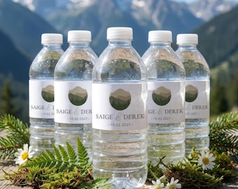 Op de afbeelding: Een verzameling heldere waterflessen met witte etiketten met een bergontwerp en de namen "SAIGE & DEREK" en de datum "10.02.2027". De flessen staan op een houten oppervlak met groen en bloemen, met een berglandschap op de achtergrond.